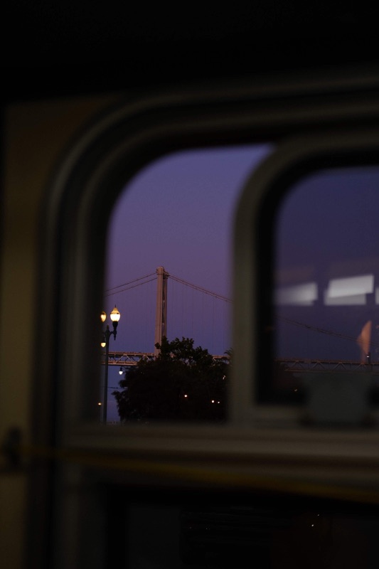 A suspension bridge and glowing street lamps at dusk, framed through a dark window.