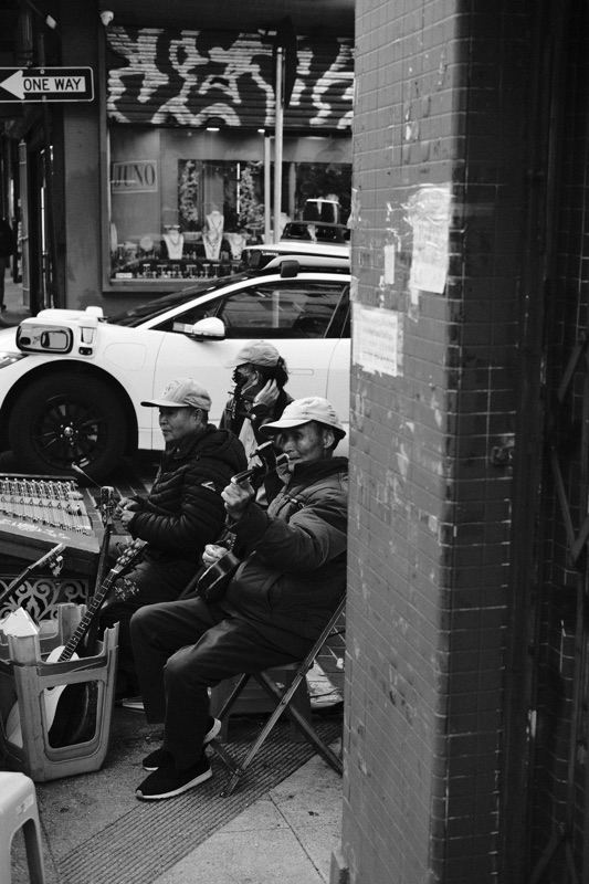 A black-and-white street scene of seated musicians beside a brick wall, with a parked car behind them.