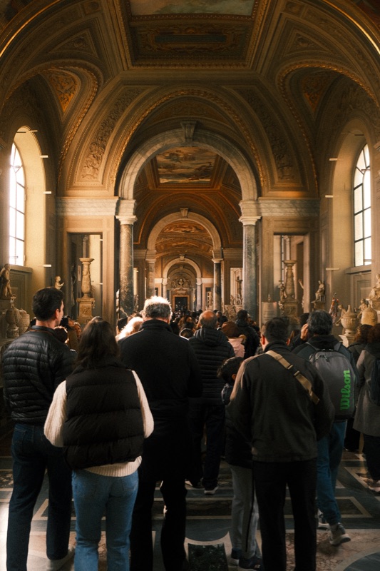 Museum visitors walking beneath an ornate vaulted ceiling lit by tall windows.