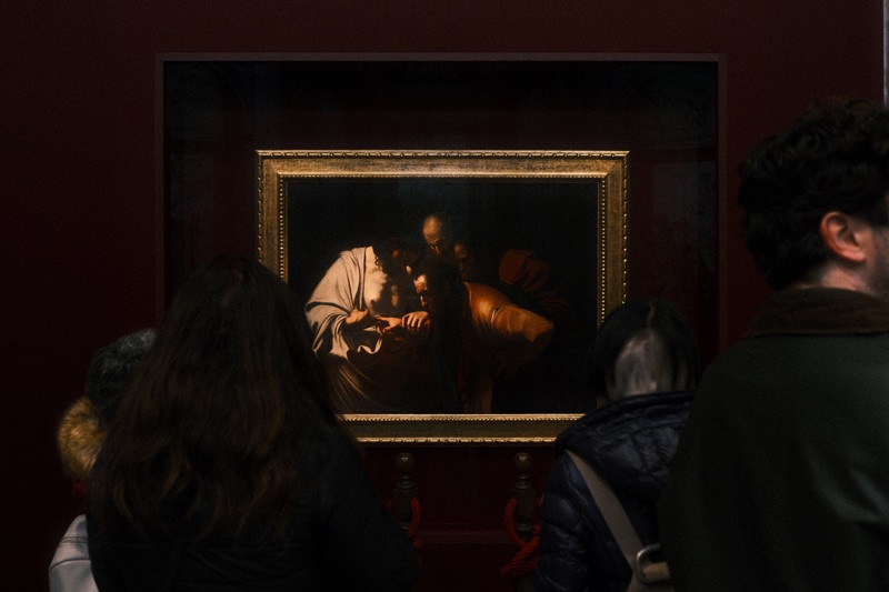 Museum visitors studying a warmly lit painting in a dark red gallery.